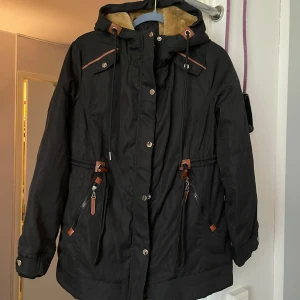 Svart parkajacka med teddyfoder - Svart parkajacka med mysigt beige teddyfoder och snygga bruna detaljer. Jackan har huva, snörning i midjan, dragkedja och knappar framtill samt två fickor med dragkedja. Perfekt för kalla dagar och har en chill, urban vibe.helt ny oanvänd 