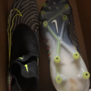 Nike fotbollsskor med neon-detaljer - Säljer ett par grymma Nike fotbollsskor med svart och grått mönster, samt neon-gula detaljer och logga. Skorna har en stickad ovandel och plös, samt platt sula med dobbar för bästa grepp på planen. Perfekt för dig som vill sticka ut på matchen!