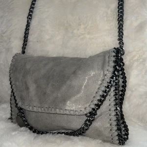 Grå axelväska med kedjedetaljer - Snygg grå axelväska med glansig yta och grova kedjor längs kanterna och axelremmen. Insidan har ett leopardmönstrat foder i brunt och svart samt praktiska fack med dragkedja. Perfekt för dig som gillar att sticka ut med coola detaljer. Säljer då denna väska inte kommer till användning. 