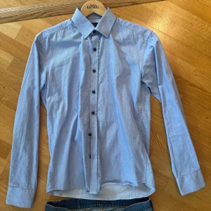 Blå slim fit skjorta från Selected Homme - Snygg blå skjorta från Selected Homme i slim fit-modell. Skjortan har diskret randigt mönster, klassisk krage och svarta knappar framtill. Tillverkad i bomull och är lätt att stryka. Perfekt för dig som gillar stilrena och moderna plagg.