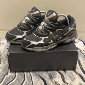 Svarta och silver Asics sneakers - Säljer ett par snygga Asics sneakers i svart och silver med coola detaljer. Skorna har mesh- och syntetpaneler, rund tå och platt sula. De har snörning och en chunky design som ger en sportig vibe. Perfekta för dig som gillar streetstyle och komfort.