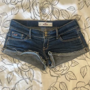 Mörkblå jeansshorts från Hollister - Snygga mörkblå low waist jeansshorts från Hollister med fransig och uppvikt kant. Klassisk femficksmodell med dubbla knappar framtill och detaljerade sömmar på bakfickorna. Perfekta för varma dagar och har en cool, avslappnad vibe.