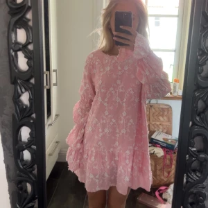 Rosa blommig kortklänning med volang - (Säljs ej rosa, den är färgad❣️)Charmig rosa klänning med blommigt mönster och volangdetaljer. Den har långa ärmar och en öppen rygg med knytning. Perfekt för en somrig look! 