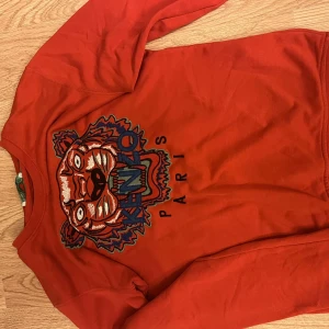 Röd Kenzo sweatshirt - Säljer en röd sweatshirt från Kenzo i storlek S med det ikoniska broderade tigermotivet och Kenzo Paris-text på bröstet. Tröjan har rund hals och långa ärmar, tillverkad i mjuk bomull. Perfekt för dig som vill sticka ut med en fet design.