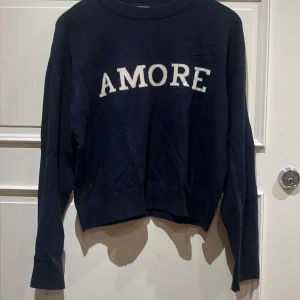 Mörkblå stickad tröja med AMORE från Gina Tricot - Snygg mörkblå stickad tröja från Gina Tricot i storlek 158/164. Framsidan har ett stort vitt AMORE-tryck i fluffigt material. Tröjan har rund hals, långa ärmar och en loose passform. Perfekt för chill dagar och enkel att matcha med jeans eller leggings.