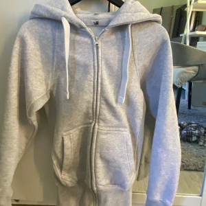 Ljusgrå ziphoodie Russemerch - Ljusgrå hoodie från Russedress i storlek XXS. Tröjan har huva med snörning, dragkedja framtill och två stora fickor. Mjuk bomullskänsla och ribbade muddar vid ärmar och nederkant. Perfekt för hösten och vintern.