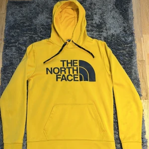 Gul hoodie från The North Face - Säljer en gul hoodie från The North Face med stor svart logga på bröstet. Tröjan har huva med svarta snören, känguruficka och långa ärmar. Materialet är mjuk polyester som känns skönt mot huden. Perfekt för chill dagar eller när du vill sticka ut med färg.