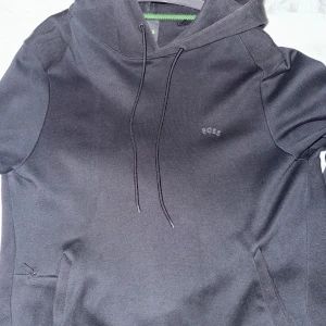 Svart hoodie från BOSS med huva - Säljer en stilren svart hoodie från BOSS med diskret logga på bröstet. Tröjan har huva med snörning och praktiska fickor på sidorna. Perfekt för dig som gillar enkel och snygg streetwear. Passar dig som vill ha en bekväm och cool look.