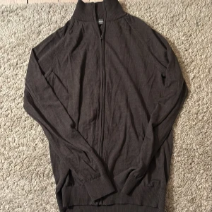 Mörkbrun kofta från Dressmann i merinoull - Säljer en stilren mörkbrun kofta från Dressmann i mjuk merinoull. Tröjan har dragkedja framtill, ribbad krage och muddar vid ärmslut och nederkant. Perfekt för dig som gillar klassisk och enkel design med skön passform.