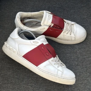 Valentino Garavani Open sneakers vit/röd - Säljer ett par Valentino Garavani Open sneakers i vitt skinn med bred röd läderpanel över mitten. Klassisk rund tå, platt sula och snörning framtill. Snygga nitar längs hälen ger extra edge. Perfekt för dig som gillar lyxiga och stilrena sneakers. Priset ej hugget I sten.