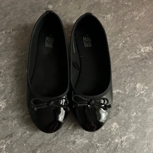 Svarta ballerinaskor med rosett - Snygga svarta ballerinaskor från Minitude med glansig finish och rund tå. Skorna har en söt rosett framtill och en klassisk, stilren look som passar till många outfits. Perfekt för dig som gillar enkel och chic stil.