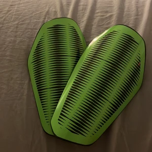 Gröna benskydd med svart mönster - Säljer ett par benskydd i cool grön färg med svart grafiskt mönster på utsidan. Insidan är svart med hål för ventilation. De är tillverkade i slitstark plast och har en modern, sportig form som passar perfekt för fotbollsträning och match. Inte använd mycket alls. Passar på en 13-åring 