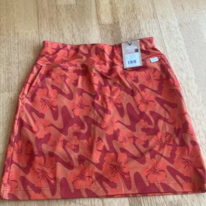 Orange blommig skort från Skhoop - Snygg orange skort från Skhoop i modellen Gudrun Skort, med blommigt mönster och inbyggda grå shorts under. Kjolen har en bekväm midja och är perfekt för aktiva dagar. Materialet är mjukt och följsamt, och färgen är riktigt poppande morotsorange.