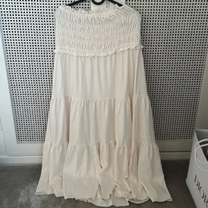 Ljus långkjol med volanger - Aldrig använd. Säljer en superfin ljus långkjol med offshoulder och smock upptill. Klänningen har flera lager med volanger och ett flowigt fall, perfekt för sommarens alla tillfällen. Färgen är mjukt beige och tyget känns lätt och luftigt.