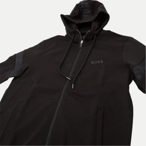 Boss kofta - Säljer en stilren svart hoodie från Boss med dragkedja och huva. Jackan har långa ärmar och ett diskret märke på bröstet. Perfekt för en casual look.