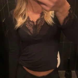 Svart långärmad topp med spets - Supersnygg svart långärmad topp med djup v-ringning och spetsdetaljer både vid halsringningen och ärmsluten. Tunn och stretchig, perfekt att styla med jeans eller kjol för en trendig look.