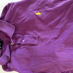 Lila Polo t-shirt från Ralph Lauren - Säljer en lila Polo t-shirt från Ralph Lauren i storlek L. Klassisk pikémodell med krage, två knappar och den ikoniska gula broderade loggan på bröstet. Perfekt för dig som gillar stilrena och färgstarka plagg.