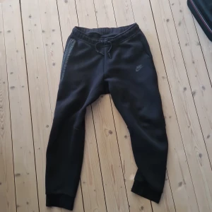 Svarta Nike tech fleece byxor XL kids - Svarta mjukisbyxor från Nike i storlek XL, med diskret Nike-logga på benet. Byxorna har snörning i midjan, ribbade muddar vid benslut och dragkedja på fickan. Perfekta för chill eller träning, tillverkade i mjukt bomullsmaterial. Andvänd typ 5 gånger.