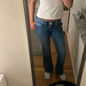 Blå bootcut jeans från Gina Tricot - Snygga blå jeans från Gina Tricot i bootcut-modell med låg midja. Jeansen har kontrastsömmar, klassiska fickor och en cool knappdetalj framtill. Perfekta för dig som gillar en avslappnad och trendig stil. Materialet är klassisk jeans i bomull.
