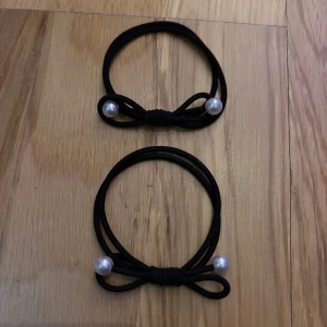 Svarta armband med pärldetaljer - Två svarta armband med elastisk design och dekorativa rosetter. Varje armband har två vita pärlor som ger en stilren och söt touch. Perfekt för att ge din outfit lite extra edge och passar till både jeans och klänning.