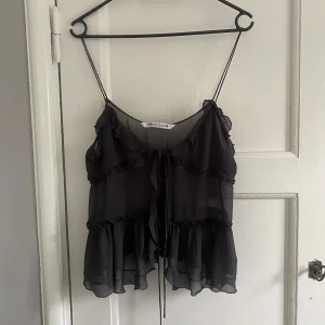 Svart volangblus från Zara - Säljer en svart blus från Zara med tunna axelband och transparent, flowy material. Blusen har volanger och dekorativa knytband framtill!