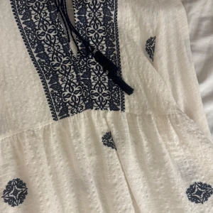 Vit och marinblå boho-blus - Supersöt vit blus med marinblå broderade detaljer och mönster. Har en v-ringad hals med snörning och tofsar, samt ett bohemiskt mönster framtill och på ärmarna. Materialet är lätt och luftigt, perfekt för varma dagar. Passformen är normal och blusen har lång ärm.
