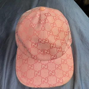 Rosa keps från Gucci med GG-mönster - Snygg rosa keps från Gucci med ikoniskt GG-mönster över hela kepsen. Justerbar rem baktill i rosa läder och diskret guldfärgad GG-detalj på sidan. Kepsen är tillverkad i bomull och polyester, perfekt för dig som vill sticka ut med en lyxig accessoar.
