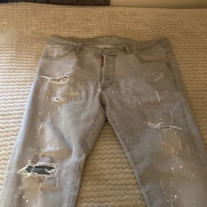 Dsquared2 ljusgrå jeans med slitningar - Säljer ett par ljusgrå jeans från Dsquared2 med coola slitningar och färgstänk på benen. Modellen har klassisk femficksdesign, rak passform och låg midja. Jeansen är tillverkade i bomull och har en streetig vibe med detaljerade trasiga partier och färgprickar.