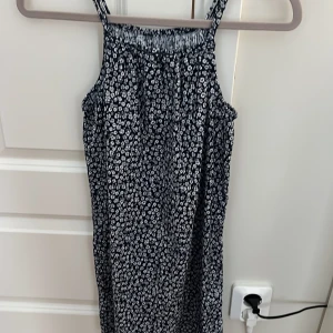 Blommig blå kortklänning från H&M - Supersöt blå kortklänning från H&M med vita små blommor över hela tyget. Klänningen har smala axelband och är ärmlös, perfekt för varma dagar. Materialet är mjukt och lätt bomull som känns skönt mot huden. Passformen är normal och klänningen har en avslappnad siluett.