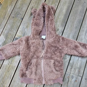 Brun teddyjacka med öron från Lindex - Supermjuk och fluffig brun teddyjacka från Lindex med huva och söta öron. Jackan har dragkedja framtill och två fickor. Perfekt för kyliga dagar när du vill vara både varm och gullig.