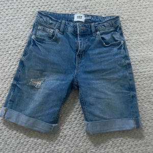Blå jeansshorts från 157, storlek 140 - Snygga blå jeansshorts från 157 i storlek 140. Klassisk femficksmodell med uppvikta benslut och slitning på ena benet för en cool look. Perfekta för varma dagar och passar dig som gillar avslappnad stil.