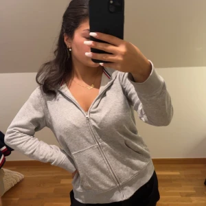 Grå hoodie från GANT med dragkedja - Grå hoodie från GANT med hel dragkedja och huva. Tröjan har ribbade muddar, fickor fram och en snygg detalj med rödblå rand på insidan av kragen. Mjuk bomullskvalitet och klassisk GANT-logga på bröstet. Perfekt för chill dagar. Har liten rosa fläck på bröstet. 