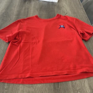 Röd t-shirt Axel Arigato S - Snygg röd t-shirt från Axel Arigato i storlek S. Modellen är croppad med korta ärmar och har en liten broderad fågel i blått på bröstet. Tillverkad i mjuk 100% bomull för skön känsla. Perfekt för dig som gillar stilrena plagg med en twist.