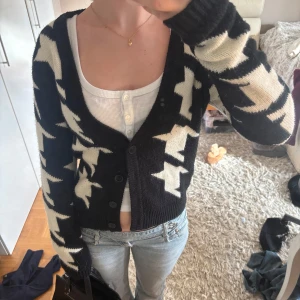 Svart och vit kofta med stjärnmönster - Säljer en svart kofta med vita stjärnor, med djup v-ringning. Koftan har långa ärmar och knäpps med svarta knappar framtill. Materialet är stickat och känns mjukt mot huden, perfekt att slänga över en topp för en cool look.