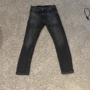 Svarta skinny jeans från Nudie Jeans - Säljer ett par svarta skinny jeans från Nudie Jeans i storlek 29/30. Jeansen har klassisk femficksdesign, svarta broderade vågor på bakfickorna och en mörk tvätt med lätt slitning. Materialet är mjukt och stretchigt denim för skön passform.
