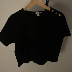 Svart croppad topp med guldknappar H&M - Svart croppad topp från H&M med korta ärmar och tre snygga guldknappar på axeln. Tjockare, lite ruffigt material som ger en cool textur. Perfekt för dig som gillar stilrena detaljer och vill ha något som sticker ut lite extra.
