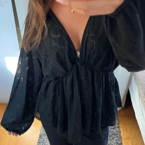 Svart blus med v-ringning och volang - Säljer en svart blus med djup v-ringning och volang nedtill. Blusen har långa, luftiga ärmar och ett lätt transparent tyg med diskret mönster. Perfekt för dig som gillar en flowy och chic stil.  Säljer då den är för stor. Bara använd en gång.
