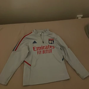 Olympique Lyonnais långärmad träningströja - Snygg långärmad fotbollströja från Olympique Lyonnais i ljusgrått med röda och blå detaljer längs ärmarna. Adidas-logga och klubbmärke på bröstet, samt 'Emirates Fly Better' tryckt framtill. Tröjan har halv dragkedja och är tillverkad i funktionsmaterial (Aeroready).