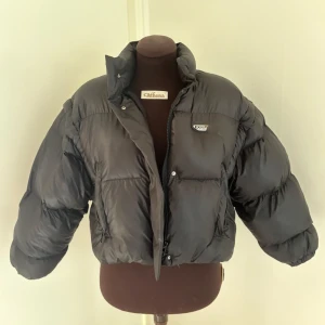 Svart croppad pufferjacka  - Svart croppad pufferjacka med hög krage och dragkedja framtill. Jackan har en quiltad look, två fickor och en snygg logga på bröstet. Perfekt för dig som vill ha en cool och trendig vinterjacka med oversized känsla.