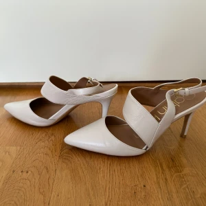 Vita pumps från Calvin Klein - Snygga vita pumps från Calvin Klein med spetsig tå och hög klack. Skorna har breda remmar över foten och justerbart spänne vid hälen. Tillverkade i skinn för en clean och stilren look, perfekta för att lyfta din outfit.
