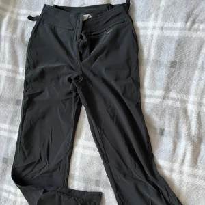 Svarta täckbyxor från Nike - Svarta täckbyxor från Nike med justerbara spännen i midjan och dragkedjor med tryckknappar vid bensluten. Byxorna har en sportig look och är tillverkade i ett slitstarkt syntetmaterial, perfekta för kalla dagar utomhus.