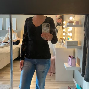 Svart spetsblus från Gina Tricot - Säljer en svart långärmad blus från Gina Tricot i genombruten spets med v-ringning och vågig kant. Perfekt för dig som gillar en lite mer edgy och stilren look. Snygg att styla med jeans eller kjol för en trendig outfit.