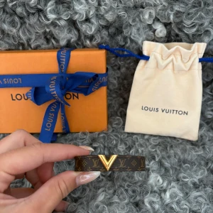 Louis Vuitton brunt läderarmband - Snyggt armband från Louis Vuitton i brunt läder med klassiskt monogrammönster och guldfärgad V-detalj framtill. Justerbar längd med guldfärgade nitar och präglad logga på insidan. Kommer med original ask och dustbag.