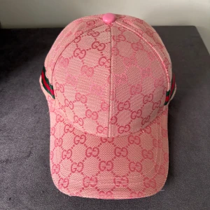 Rosa Gucci keps med GG-mönster - Snygg rosa keps från Gucci med ikoniskt GG-mönster över hela kepsen och klassiska gröna och röda ränder på sidan. Justerbar rem bak i rosa läder med Gucci-logga. Tillverkad i Italien, materialet är bomull och polyester.