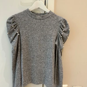 Zara Grå stickad tröja med pärlor - Supersnygg grå stickad tröja med puffiga långärmar dekorerade med pärlor längs rynkningen. Tröjan har en rund hals och mjukt material som känns skönt mot huden. Perfekt för dig som gillar stilrena detaljer och vill ha något extra till din outfit.