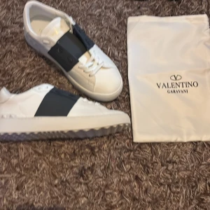 Valentino Garavani Open sneakers vit/svart - Snygga Valentino Garavani Open sneakers i vitt skinn med bred svart rem över vristen. Klassisk rund tå, platt sula med mönster och vita snören. Kommer med original dustbag. Perfekt för dig som gillar stilrena designer och exklusiva märken.