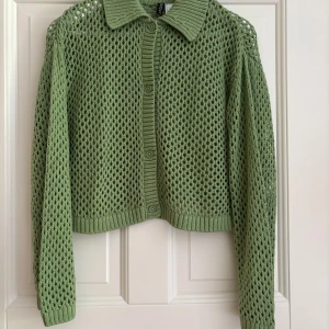 Grön stickad kofta från H&M Divided - Säljer en ljusgrön stickad kofta från H&M Divided i storlek S. Koftan har ett coolt hålmönster, ribbad krage och muddar samt knappar framtill. Perfekt för lager-på-lager och ger en chill vibe till din outfit.