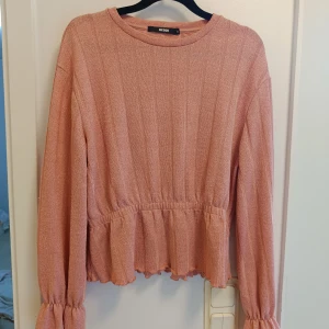 Rosa ribbad tröja med volang från Bik Bok - Supersöt ljusrosa tröja från Bik Bok med ribbad struktur och volangdetaljer vid midjan och ärmsluten. Långärmad och med rund hals, perfekt för dig som gillar en feminin och chill stil. Passar till jeans eller kjol för en snygg look.