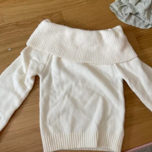 Vit offshoulder stickad tröja - Säljer en vit stickad tröja med bred ribbad offshoulder-krage. Tröjan har långärmade ärmar och ribbade muddar vid ärmslut och nederkant. Perfekt för dig som gillar en clean och stilren look med lite extra edge på halsen.