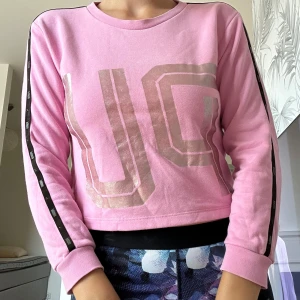 Rosa sweatshirt med silvertryck JC - Snygg rosa sweatshirt från JC med stora silverfärgade bokstäver tryckta både fram och bak. Tröjan har svarta detaljer längs ärmarna och är croppad i modellen. Mjuk bomullskvalitet och långärmad, perfekt för en chill och sportig look. Storlek 152 11-12 år (men kan användas om man har XXS/XS i vuxen storlek)